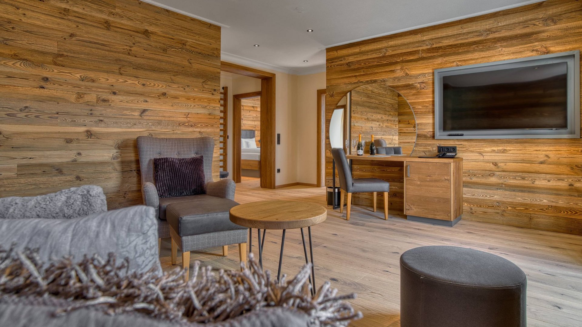 Ihr Traumurlaub im Hotel Hofbräuhaus Gemütlicher Wohnbereich einer Suite im Wellnesshotel Hofbräuhaus in Bodenmais mit moderner Couch, Sessel, Flachbild-TV, großem runden Spiegel.